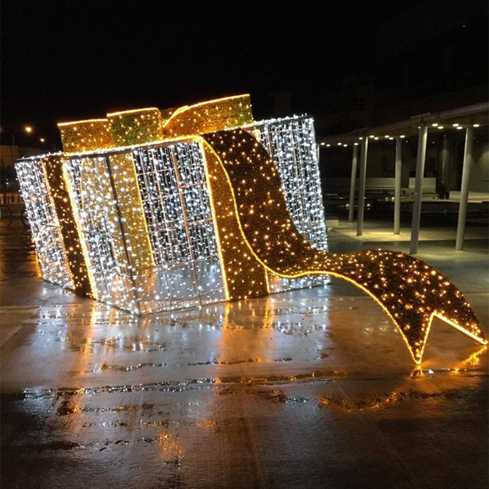 Oversized illuminated gift box with a large bow and a long decorative ribbon extending down to the ground