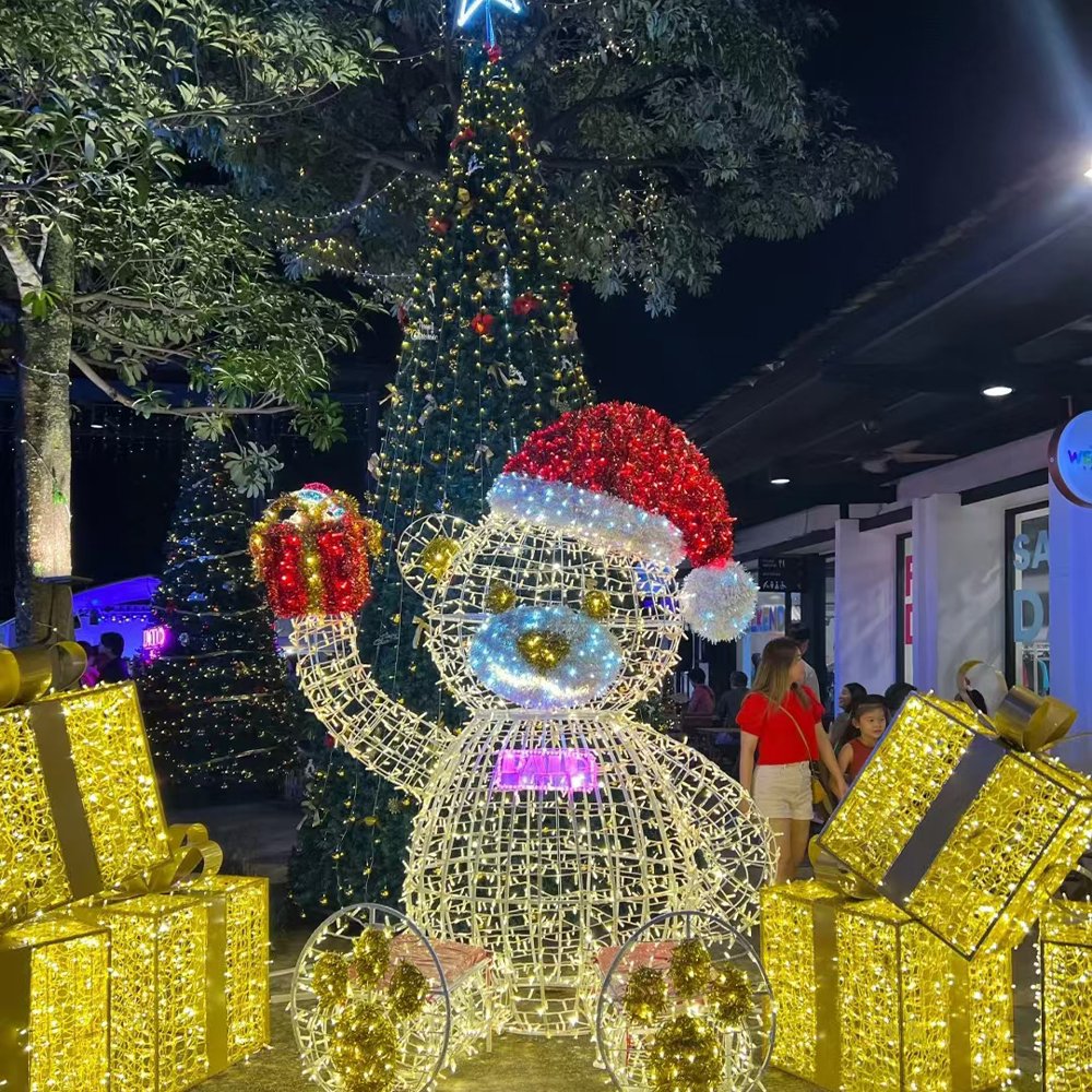 Seated illuminated teddy bear in a Santa hat holding a gift above one paw