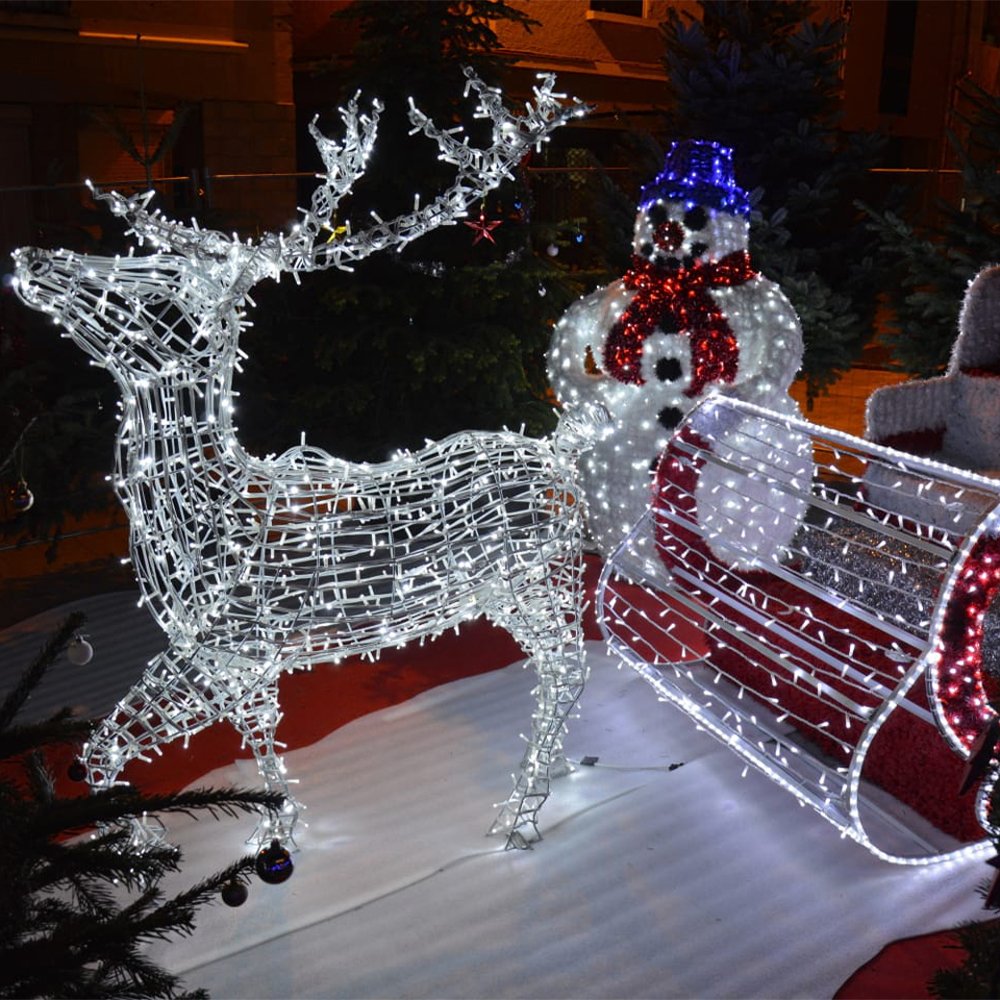 Illuminated reindeer beside a decorative sleigh and a blue-hat snowman in a Christmas display scene