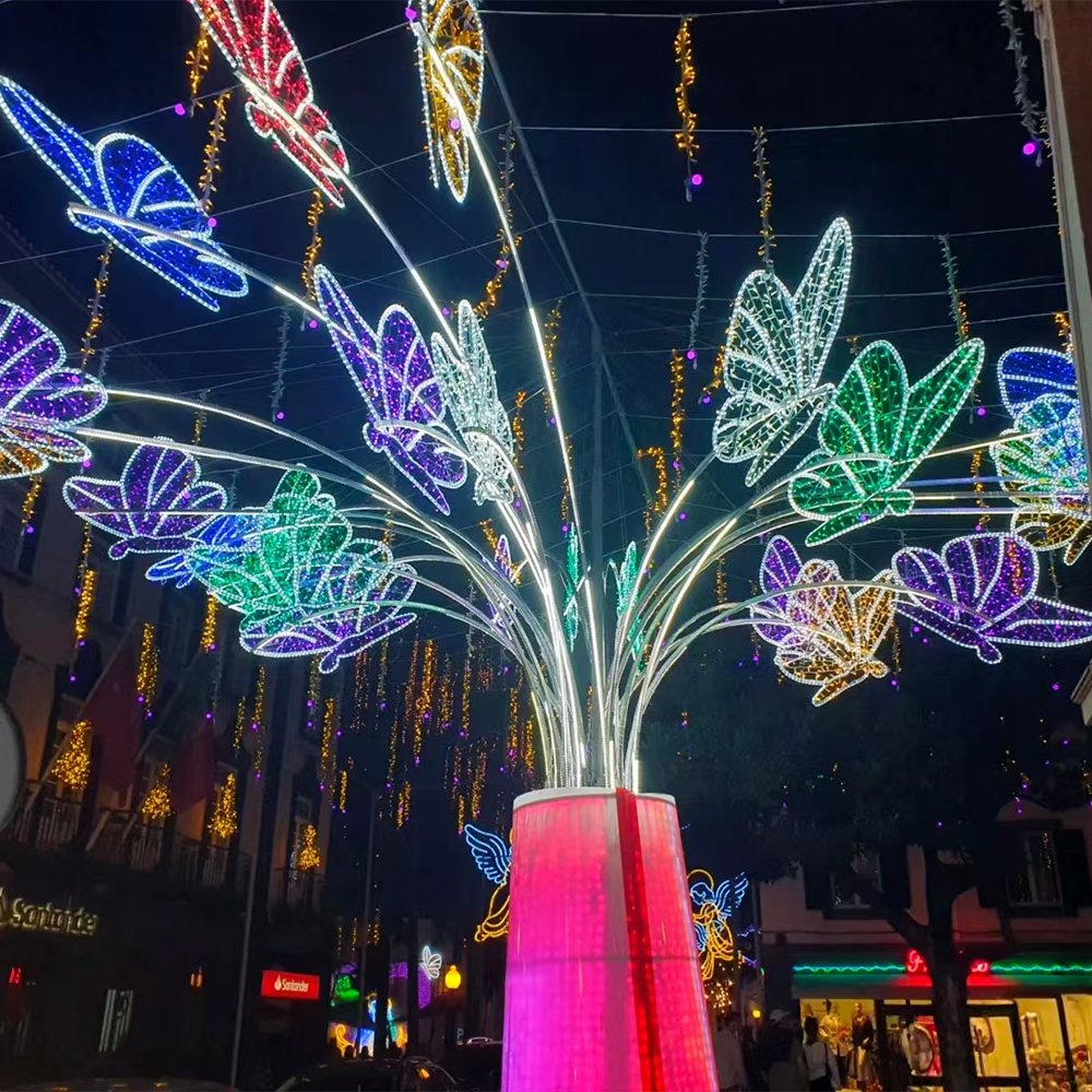 Illuminated butterfly motifs arranged on sweeping stems above a tall decorative pedestal in a city street