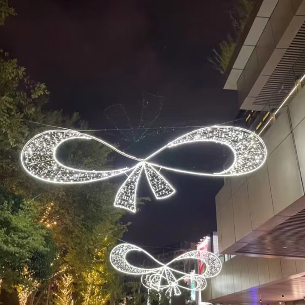 Wide white illuminated double-loop bow motif with short tails suspended beside a shopping centre