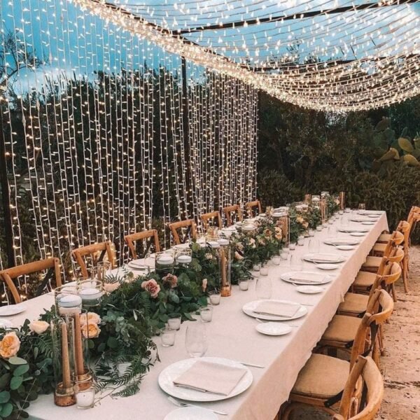 Warm white curtain lightwall beside a long outdoor banquet table with flowers and wooden chairs