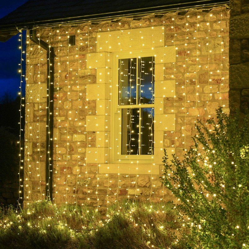 Warm white vertical curtain lights across a stone exterior wall and a pale-framed window beside garden shrubs