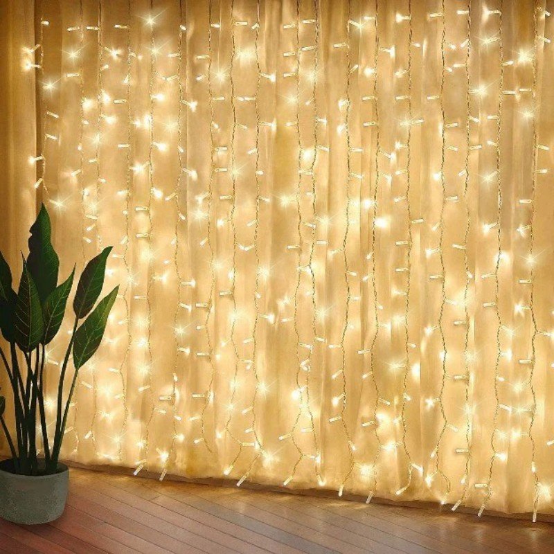 Warm white curtain LEDs hanging in front of cream fabric drapes beside a broad-leaf potted plant