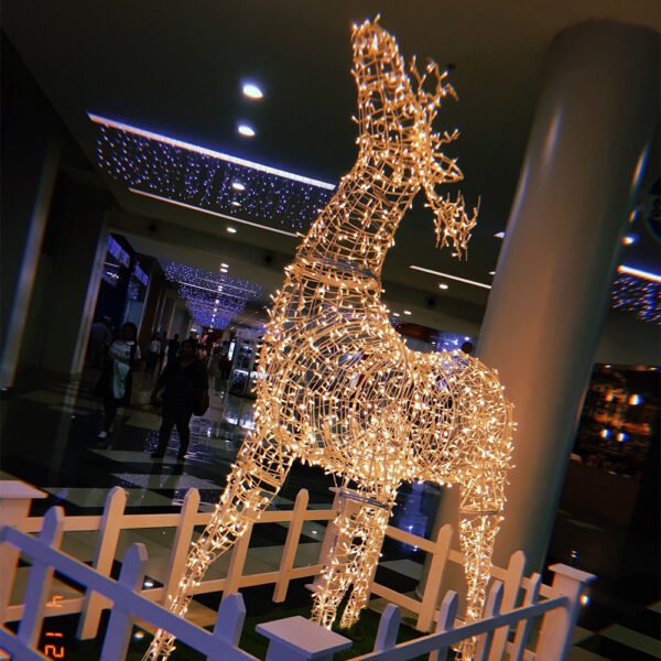 Warm-looking illuminated standing deer with an extended neck in a mall display area surrounded by a decorative low fence