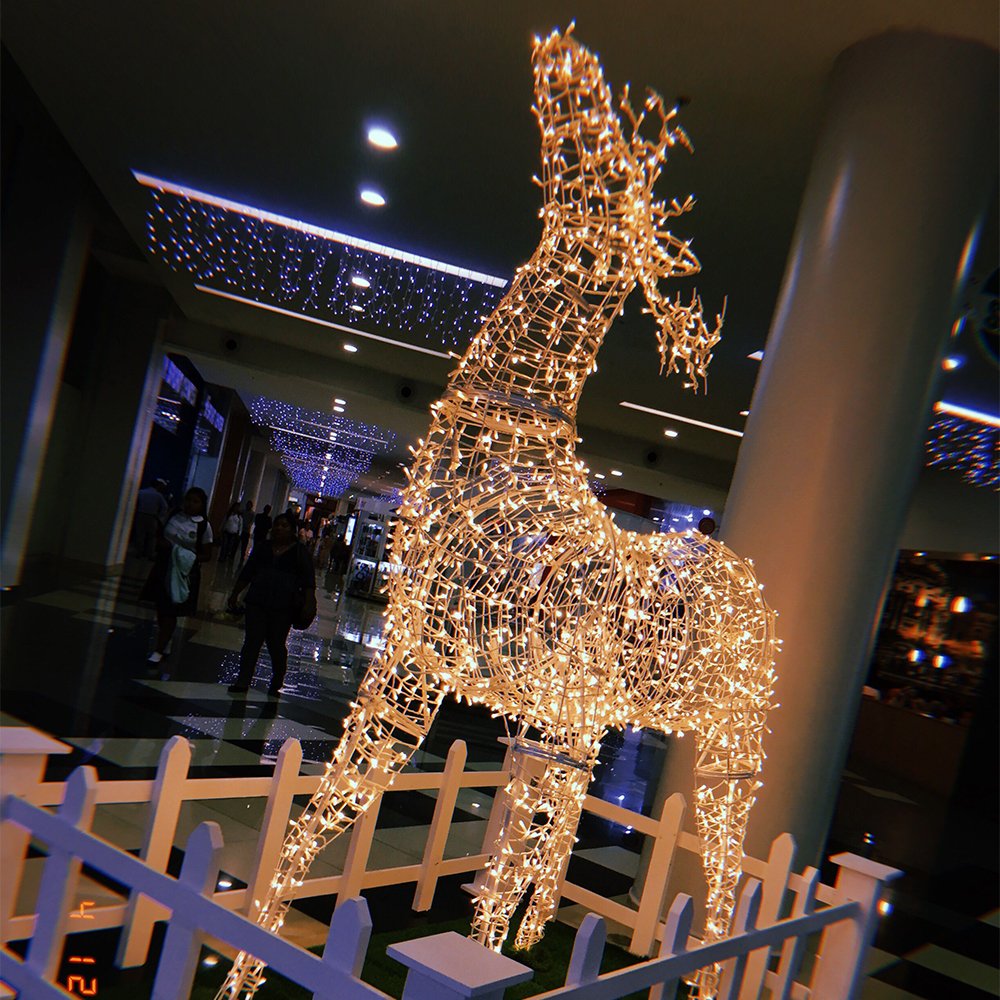 Warm-looking illuminated standing deer with an extended neck in a mall display area surrounded by a decorative low fence