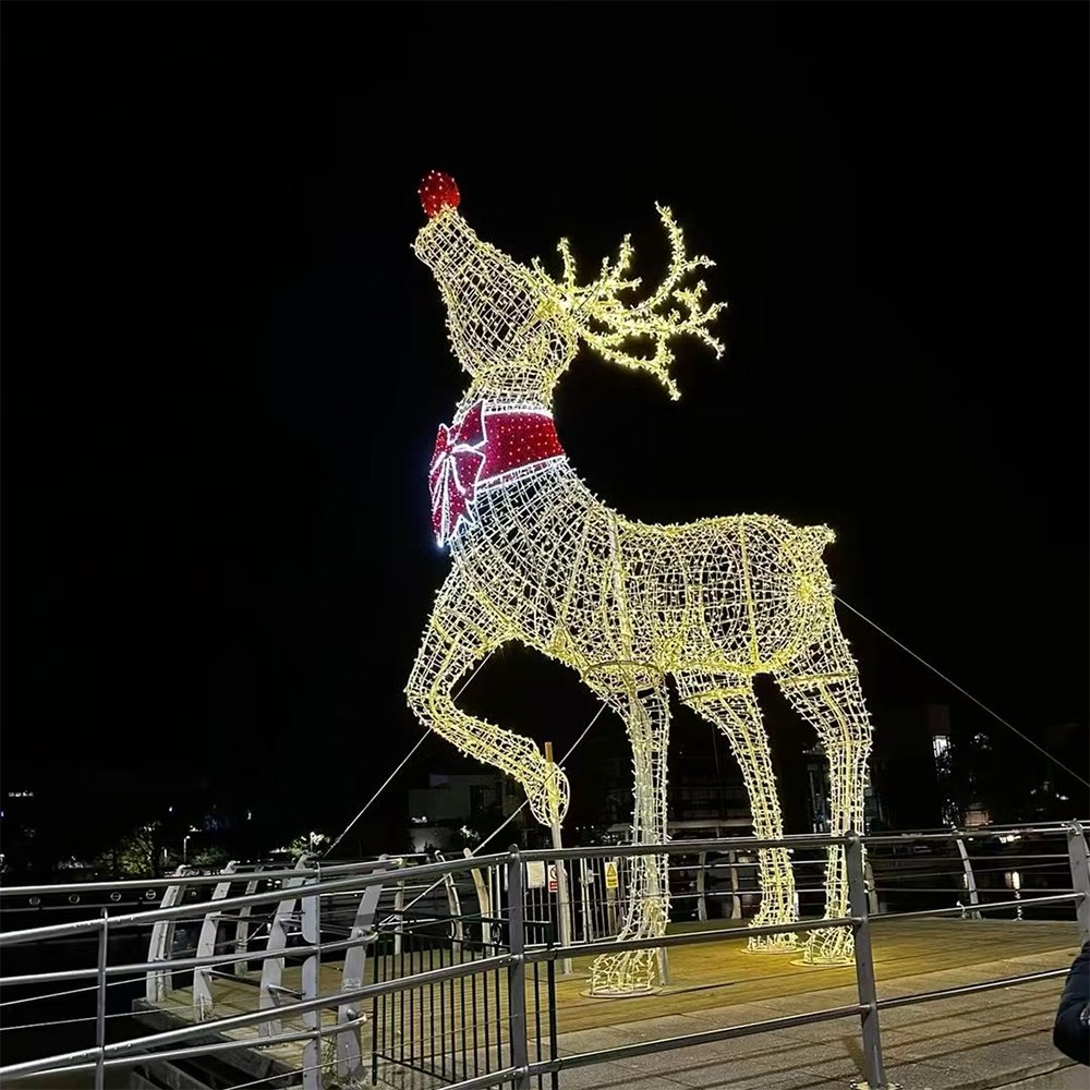 Upward-facing illuminated reindeer with branching antlers, a bow-shaped collar and one bent front leg beside a waterfront railing
