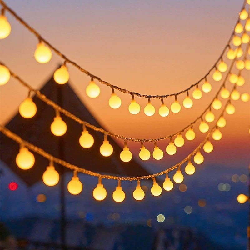 Three layered arcs of warm white globe lights against a sunset sky with a softly focused roof silhouette