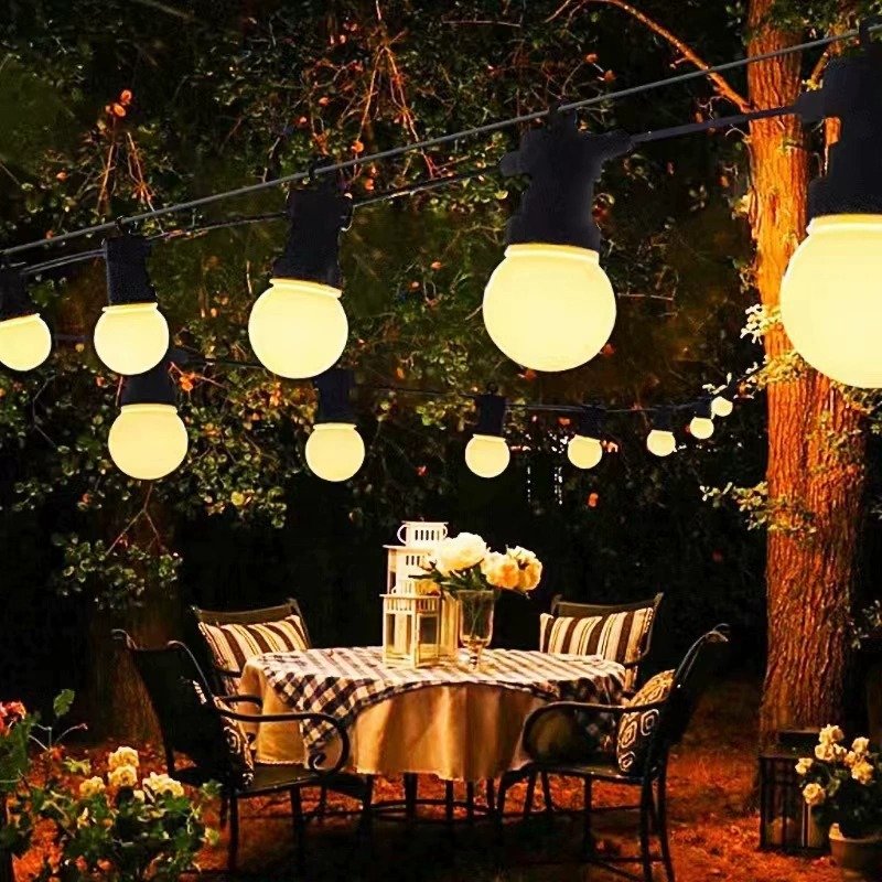 Warm white globe lights above a small garden dining table beside a tree, flowers and decorative lanterns